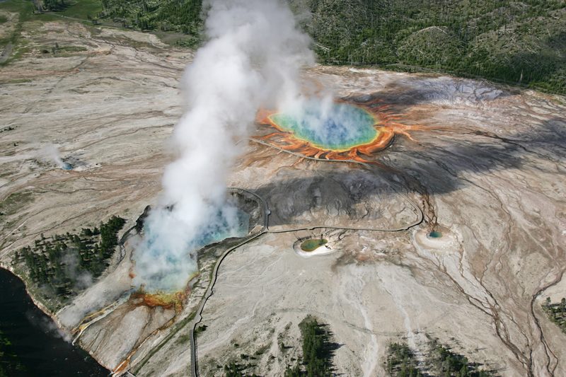Grand Prismatic Spring seen from the air, a blue pool ringed with orange and yellow mats, with steam pouring from Excelsior Geyser beside it