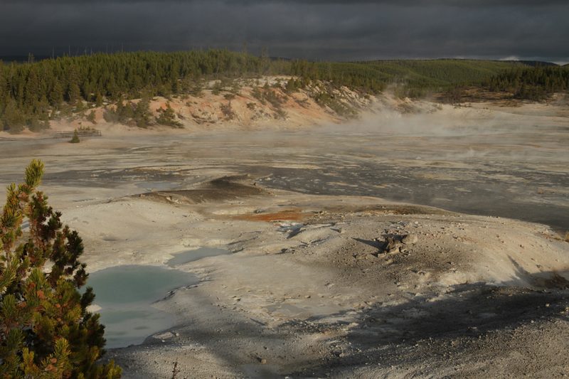 Porcelain Basin at Norris, a pale valley of bare, steaming mineral ground with a milky blue pool, under a dark sky